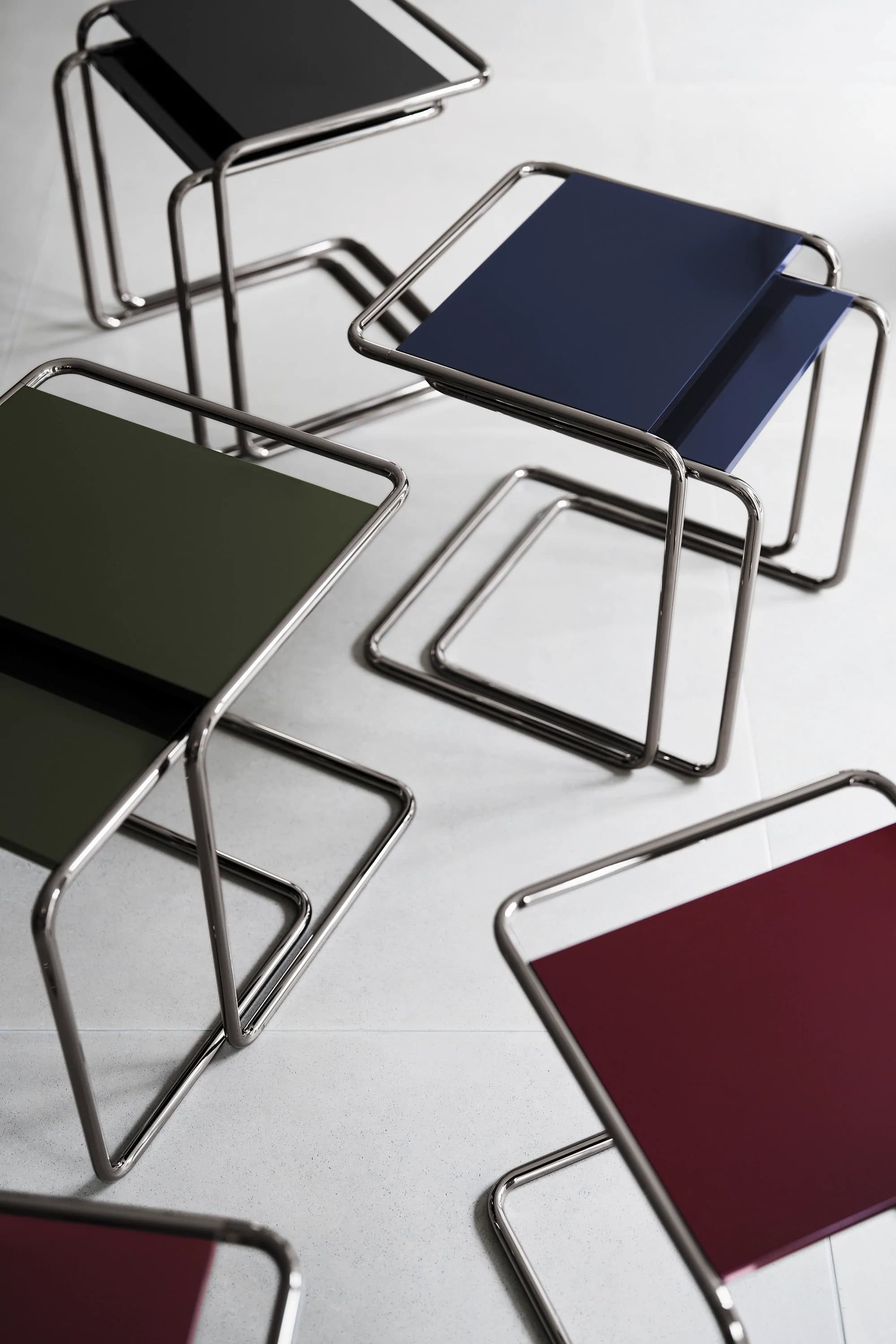 Editorial image for JS . THONET: Thonet B 97 side tables in various colors – black, dark blue, olive green and bordeaux – with chrome-plated tubular steel frame, arranged diagonally on a light background.