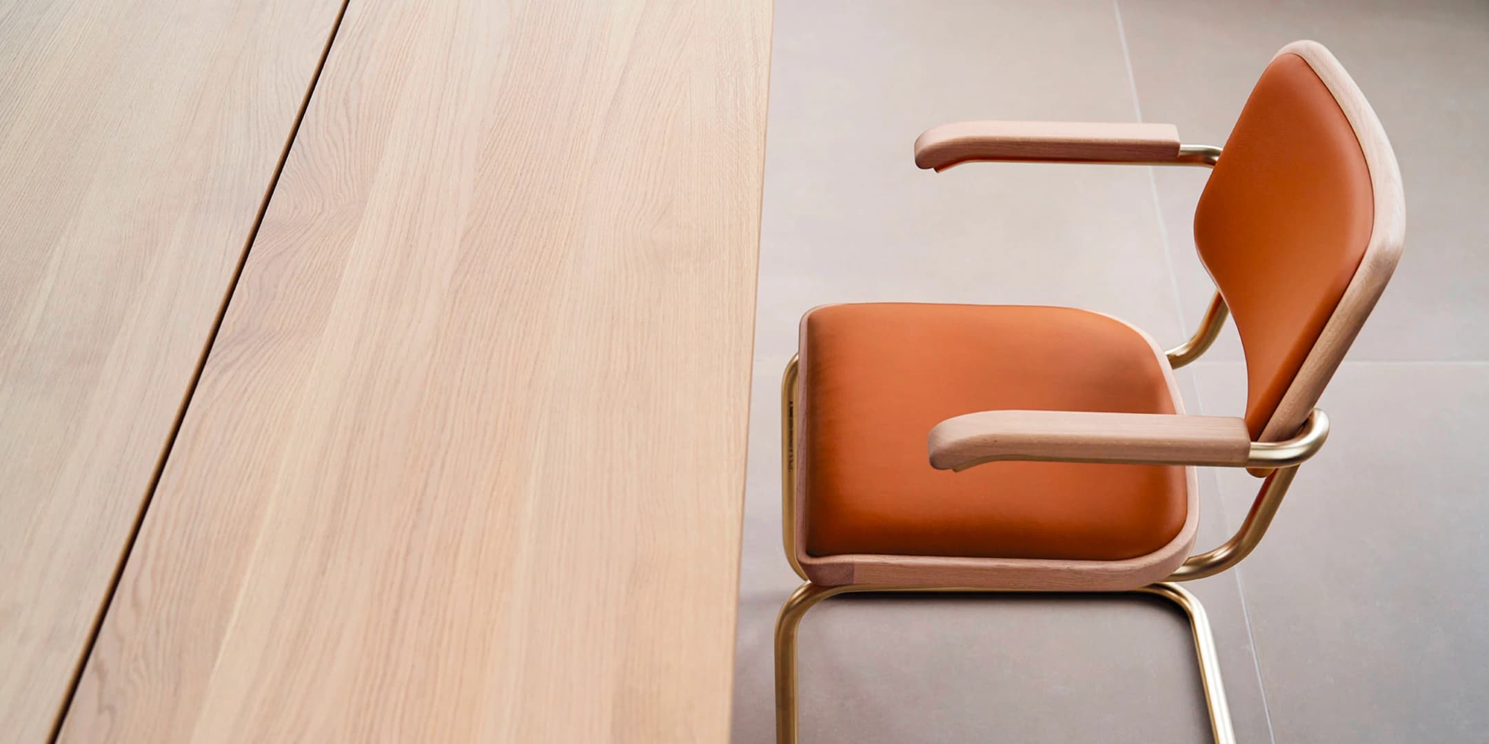Close-up of a Thonet S 64 cantilever armchair in the Nordic Edition – cognac leather, natural wooden armrests and gold-colored tubular steel frame – next to a Thonet S 1070 dining table with light oak table top.